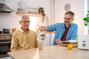 En la mesa de la cocina, un cuidador ayuda a servir una bebida a un familiar de edad avanzada.